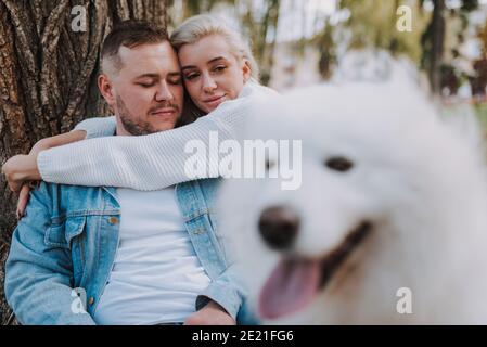 Happy husband and his wife posing with white furry playful dog samoyed breed while hugging near big tree in autumn park Stock Photo