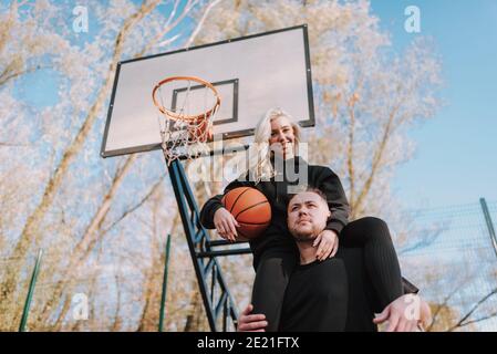 Low angle view of smiling lady and man in black sportswear posing on basketball court outdoors Stock Photo
