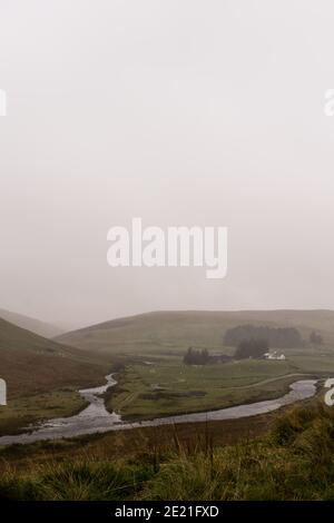 Looking through a car window blurred by raindrops. As a river meanders past a farm house on a bleak moorland valley. On a moody, misty day. Wales, UK. Stock Photo