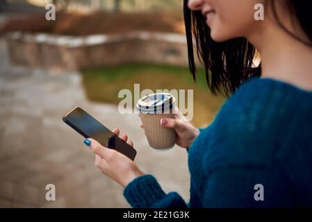 Close up of an unknown smiling female model looking on screen of a cellphone and drinking coffee outdoors Stock Photo