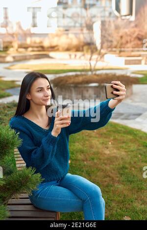 Positive female student making photo of herself on a mobile phone while relaxing with cup of coffee in a quiet corner of a picturesque square Stock Photo