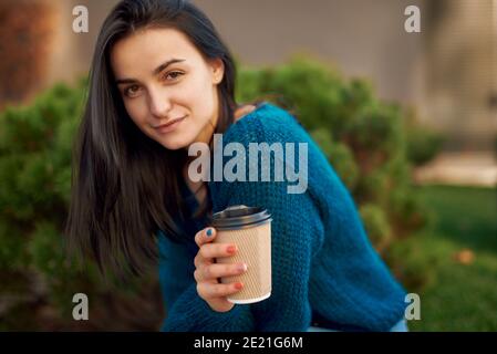 Young long-haired woman with a beautiful smile feeling content and calm relishing the moment while sitting on bench and drinking coffee Stock Photo