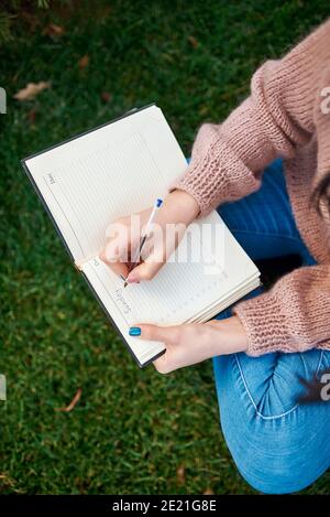 Top view of young lady in casual clothing writing something down to her daily planner while sitting cross-legged on green sward Stock Photo