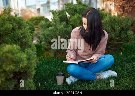 Confident female student writing messages at her daily planner and spending time outdoors in a warm day Stock Photo