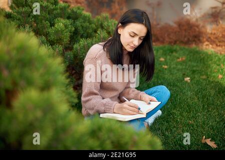 Pleasant young brunette with long hair is concentrated on writing on page of her notebook outside on an open lawn Stock Photo
