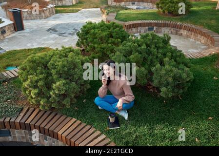 Pretty young student with a charming smile speaking by phone while spending time on a grass sward in cosy city square Stock Photo