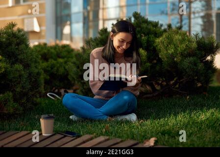 Nice-looking smiling young model posing with a book on grass in quiet and peaceful dooryard with pines outside of modern edifice Stock Photo