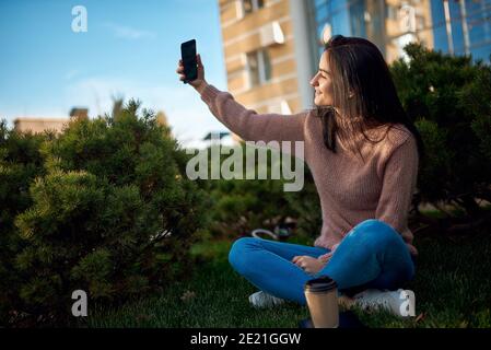 Young beautiful dark-haired woman taking heartwarming picture of herself and sitting outdoors Stock Photo