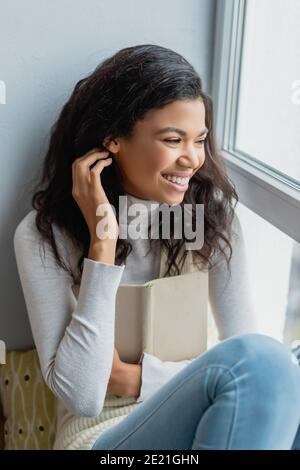 cheerful african american woman fixing hair and looking through window while holding book at home Stock Photo