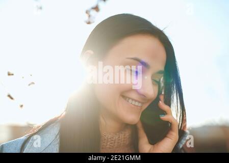 Close up shot of a nice-looking young woman with charming smile communicating with someone by phone outdoors Stock Photo