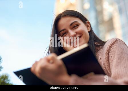 Beautiful young female looking over the pages of book and holding in hand with smile on blurred background Stock Photo