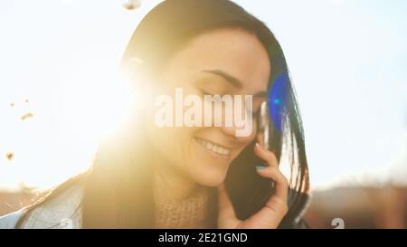 Close up portrait of an adorable young lady taking pleasure from a cheerful phone conversation and sunny autumn evening Stock Photo