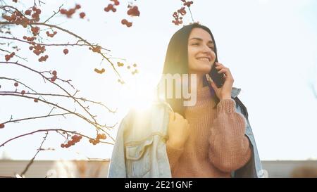 Charming young lady standing outdoors in sunlight and having pleasant conversation by cellphone Stock Photo