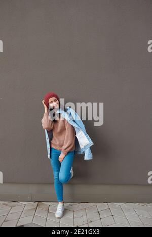 Smiling young lady in stylish spring clothes posing on one leg and touching head by hand while leaning on a large dark wall outdoors Stock Photo