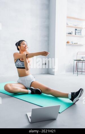happy african american woman stretching on fitness mat near laptop at home Stock Photo