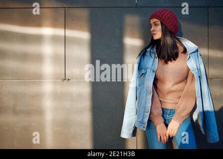 Charming slim lady in denim coat over her shoulders and knitted beanie on head standing on a street Stock Photo