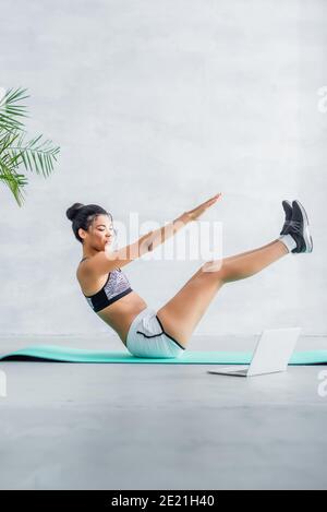 african american sportswoman exercising in boat pose near laptop at home Stock Photo