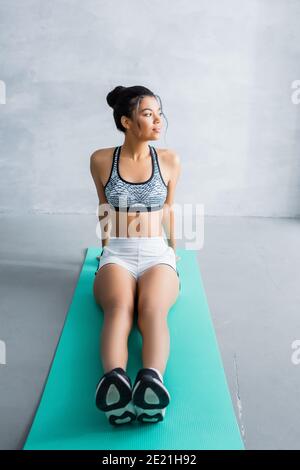 young african american woman in sportswear looking away while sitting on fitness mat at home