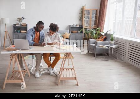 Full length portrait of caring African-American father helping son doing homework or studying while sitting together at desk in home interior, copy sp Stock Photo