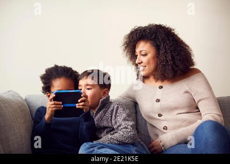 Mother And Sons Sitting On Sofa At Home Playing Computer Game Together On Hand Held Device At Home Stock Photo