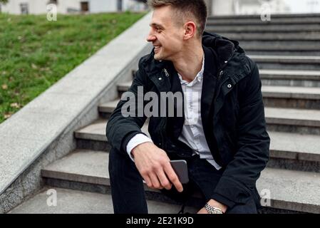 Cropped photo of smiling handsome guy resting on stairs while looking away and holding smartphone. Lifestyle concept Stock Photo