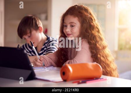 Two Children Sitting At Kitchen Counter Doing Homework Using Digital Tablet Stock Photo