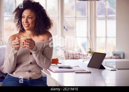 Woman With Digital Tablet Working From Home On Kitchen Counter Drinking Coffee Stock Photo
