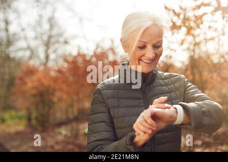 Senior Woman Running In Autumn Countryside Exercising Checking Smart Watch Fitness Activity App Stock Photo