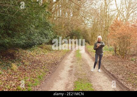 Senior Woman Running In Autumn Countryside Exercising Checking Smart Watch Fitness Activity App Stock Photo