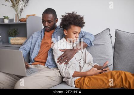African American man and his teen son sitting together on sofa in living room surfing Internet using laptop and smartphone Stock Photo