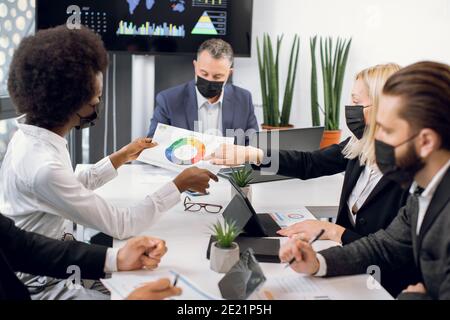 Multiracial high-skilled diverse businesspeople in black face masks, sitting around boardroom table and discussing business strategy. African and Stock Photo