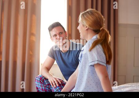 Smiling Couple Sitting On End Of Bed At Home Wearing Pajamas Stock Photo