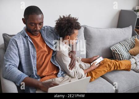 High angle shot of modern African American man and his teen son sitting relaxed together on sofa in living room using laptop and smart phone Stock Photo