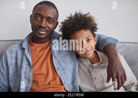 Horizontal waist up portrait shot of happy African American man sitting on sofa with his teen son looking at camera Stock Photo