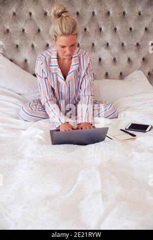 Businesswoman In Pyjamas Sitting On Bed With Laptop Working From Home During Pandemic Lockdown Stock Photo