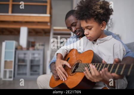 Mature man spending spare time with his son at home, teaching him how to play beautiful song on guitar Stock Photo