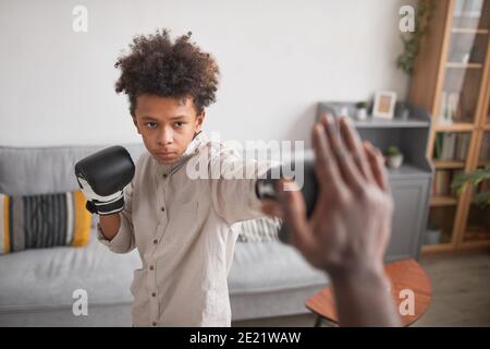 Medium portrait shot of serious African American boy wearing boxing gloves training punches with his father at home Stock Photo
