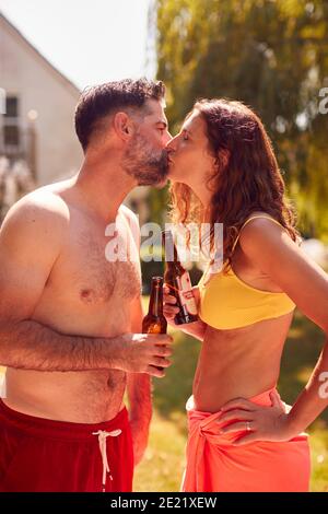 Loving Couple Wearing Swimming Costumes Standing In Pool In Summer Garden At Home Drinking Beer Stock Photo