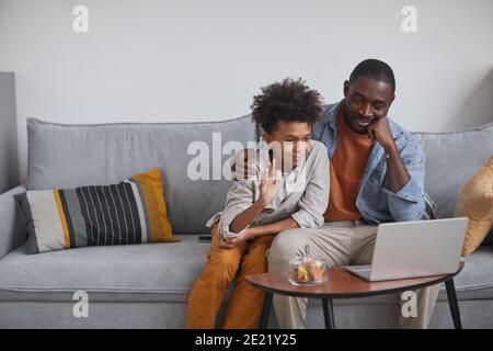 Modern father and son sitting on sofa in front of laptop screen starting video call with relatives with greeting them Stock Photo