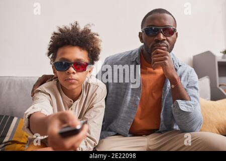 Medium portrait of stylish African American boy sitting on sofa with his father turning on 3D film using clicker Stock Photo