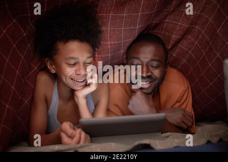 Joyful African American boy watching exciting movie on digital tablet with his father under checked blanket in darkness Stock Photo