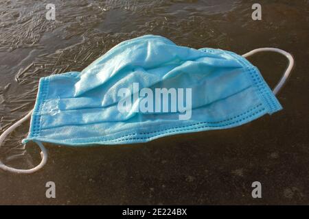 Blue protective mask on frozen river. Discarded pandemic face mask on ice in winter. Top view