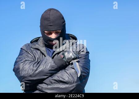 a sad, thoughtful bandit stands in a black mask and with a knife against the blue sky Stock Photo