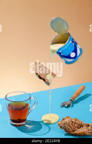 Puddle of condensed milk flows from levitating jar, can opener, black bread, glass of black tea, concept breakfast from soviet childhood, vertical, co Stock Photo
