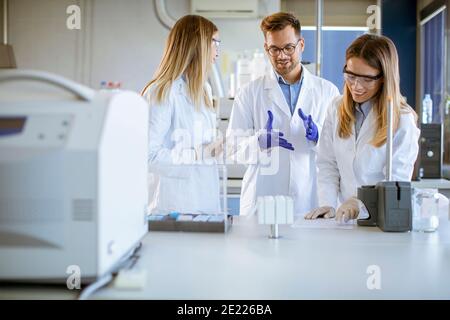 Group of young researchers in protective workwear standing in the laboratory and analyzing liquid samples at ion chromatography equipment Stock Photo