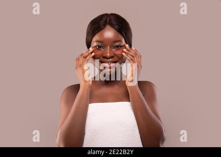 Eye skin treatment. Smiling African American woman applies golden patches under eyes, has white towel on head, watching in the mirror, in spa Stock Photo