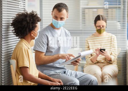 Portrait of young male doctor talking to boy waiting in line at medical clinic, both wearing masks, copy space Stock Photo