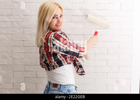 Woman painting wall in new home. Happy beautiful young woman doing wall painting. A young girl makes repairs: paints the walls with white paint using Stock Photo