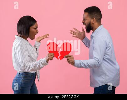 Angry young guy and lady with red heart cut in half shouting at each other, having conflict or fight on pink background Stock Photo