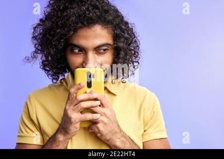 sly arabic male using smartphone, thinking what to write in message, looking at side, isolated over purple background Stock Photo